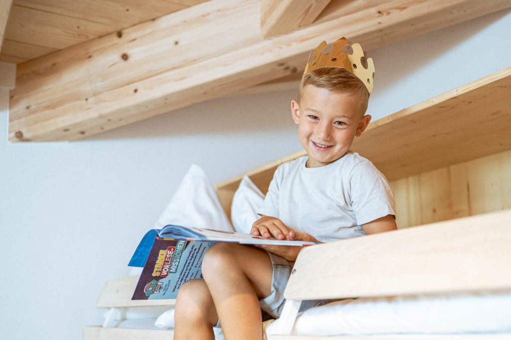 kleiner Junge mit goldener Krone sitzt am Stockbett mit Bilderbuch und lacht